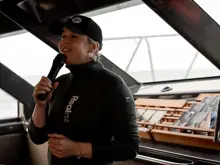 Nature guide, wearing RealNZ branded jacket and cap, talks into a microphone at the front of a catamaran cruise boat.