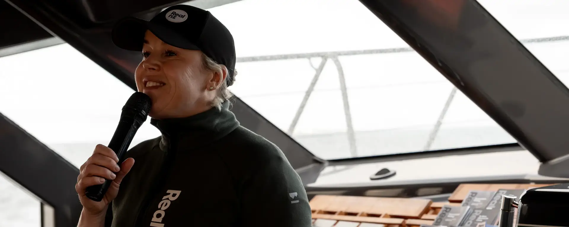 Nature guide, wearing RealNZ branded jacket and cap, talks into a microphone at the front of a catamaran cruise boat.