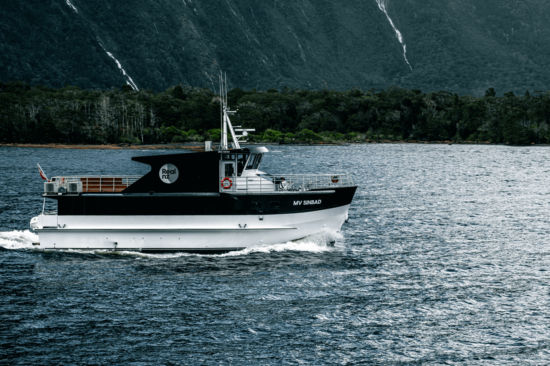A RealNZ branded small boat cruises in Milford Sound with waterfalls in the background.