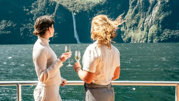 Two guests enjoying drinks on deck with Milford Sound behind them.