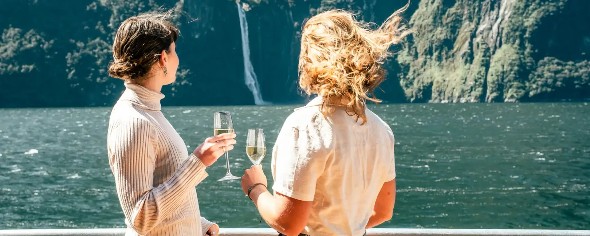 Two guests enjoying drinks on deck with Milford Sound behind them.