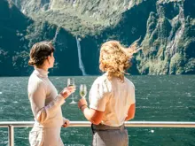 Two guests enjoying drinks on deck with Milford Sound behind them.