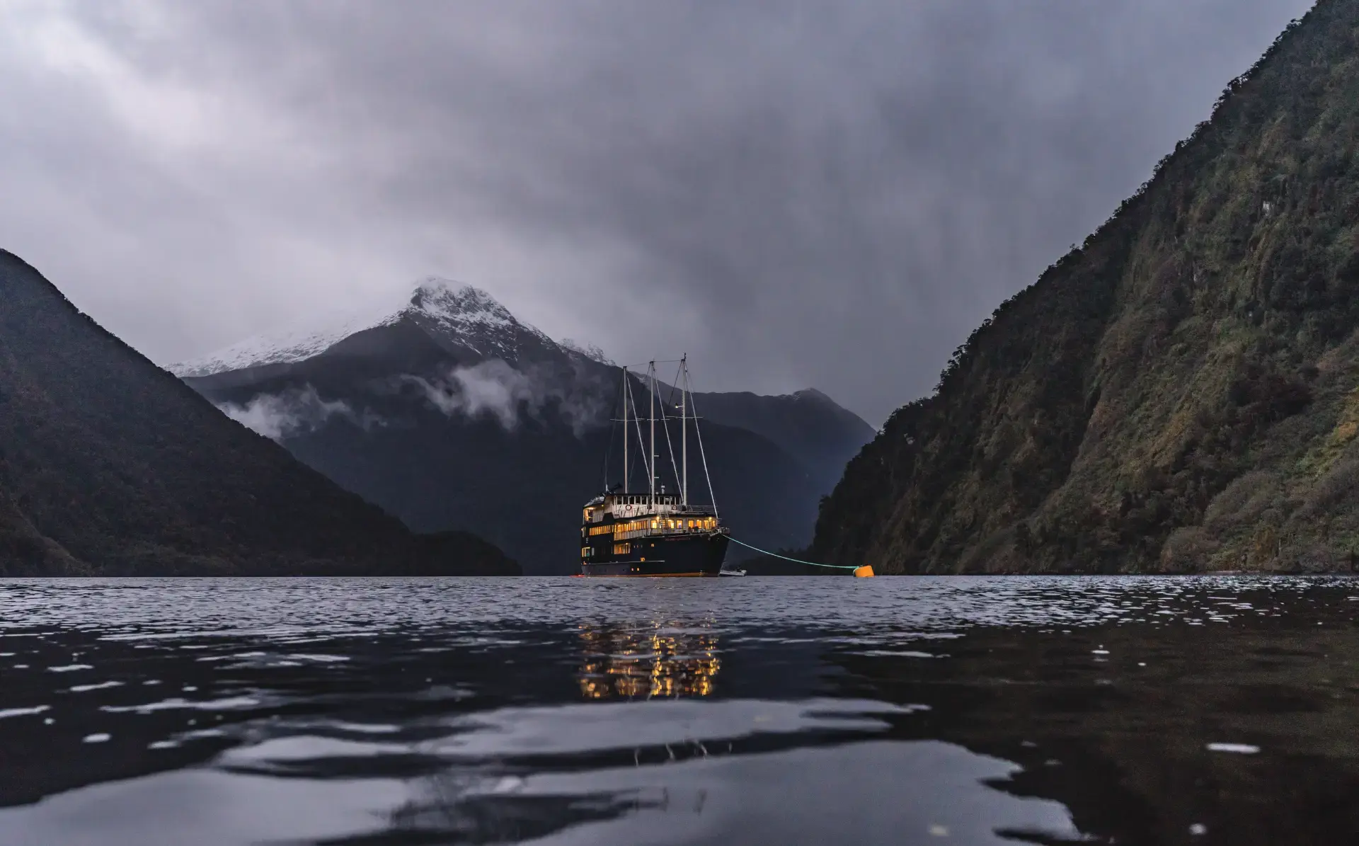 The Milford Mariner overnight cruise boat anchors in Milford Sound for the evening, surrounded by darkening waters and lit orange from the lights within. 