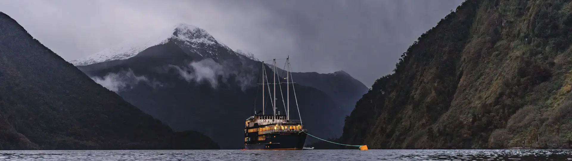 The Milford Mariner overnight cruise boat anchors in Milford Sound for the evening, surrounded by darkening waters and lit orange from the lights within. 