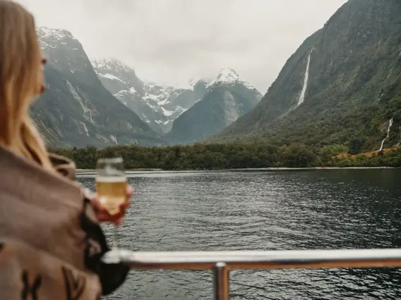 A person holding a glass of sparkling wine looks out over the calm waters of Milford Sound, surrounded by steep, misty mountains and waterfalls.