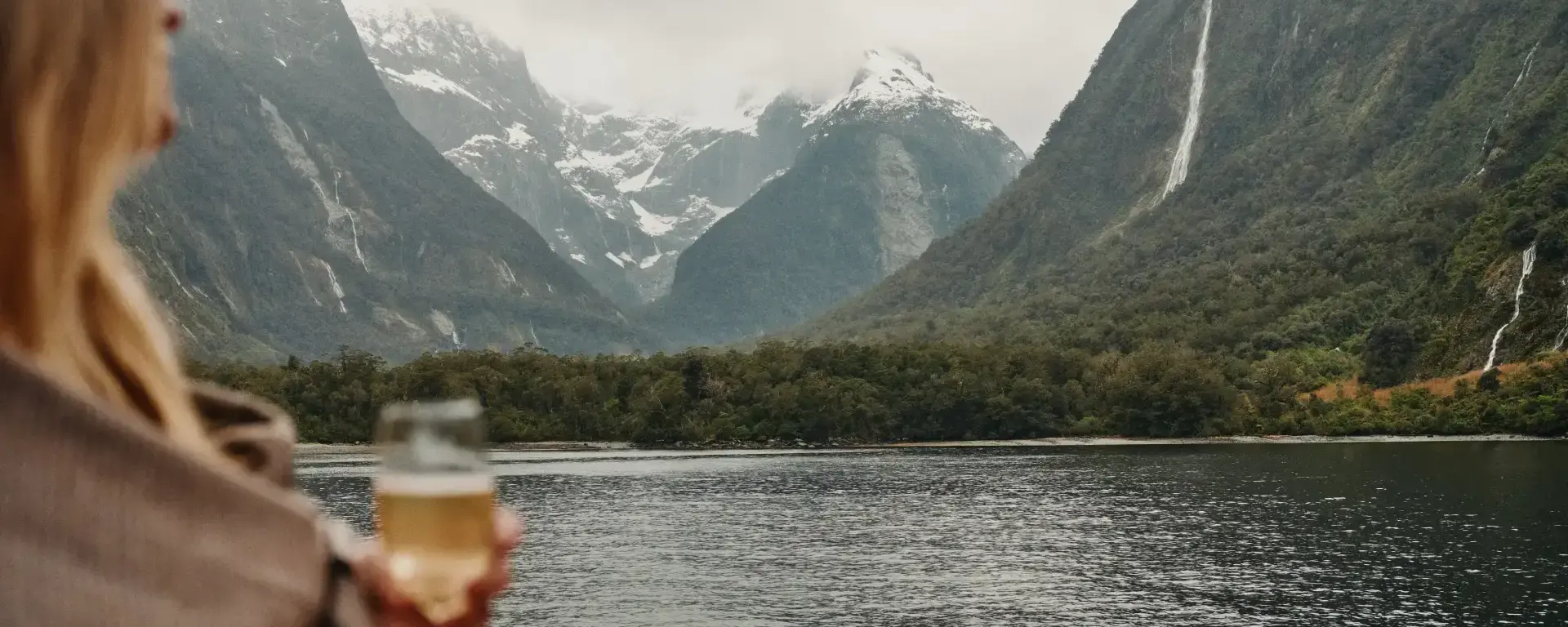 A person holding a glass of sparkling wine looks out over the calm waters of Milford Sound, surrounded by steep, misty mountains and waterfalls.