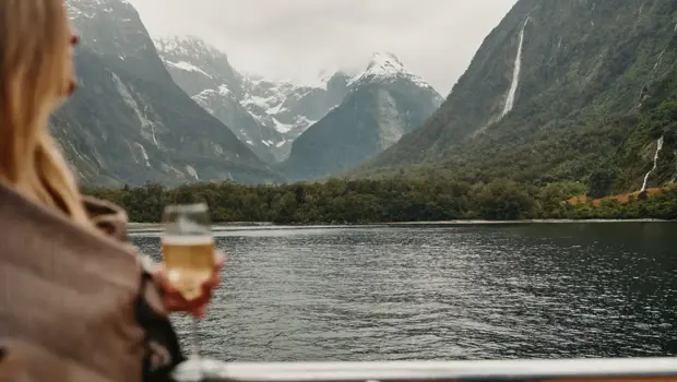 A person holding a glass of sparkling wine looks out over the calm waters of Milford Sound, surrounded by steep, misty mountains and waterfalls.