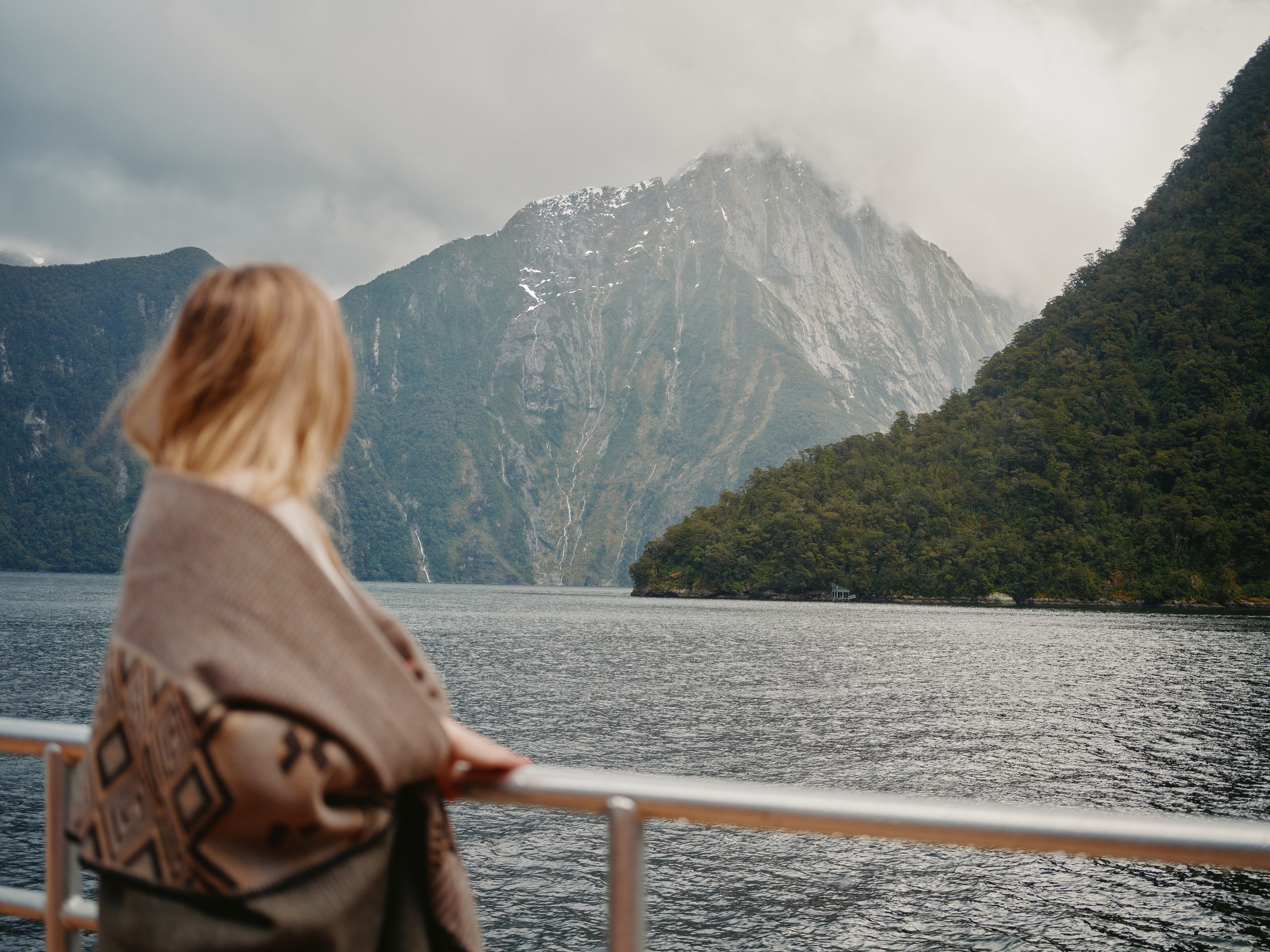 A woman standin gon the outside deck of a cruise boat with a boutique blanket wrapped around her shoulders looks out at the view of the water and surrounding mountains.