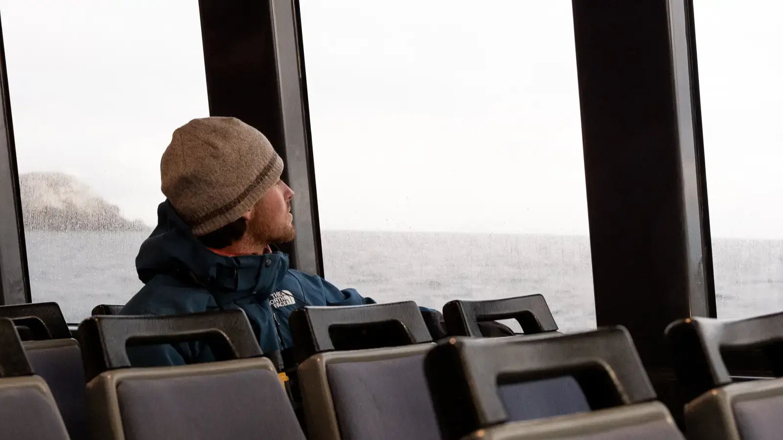 A man sits inside on a catamaran ferry as it cruises across the water, looking out the window at the view. 