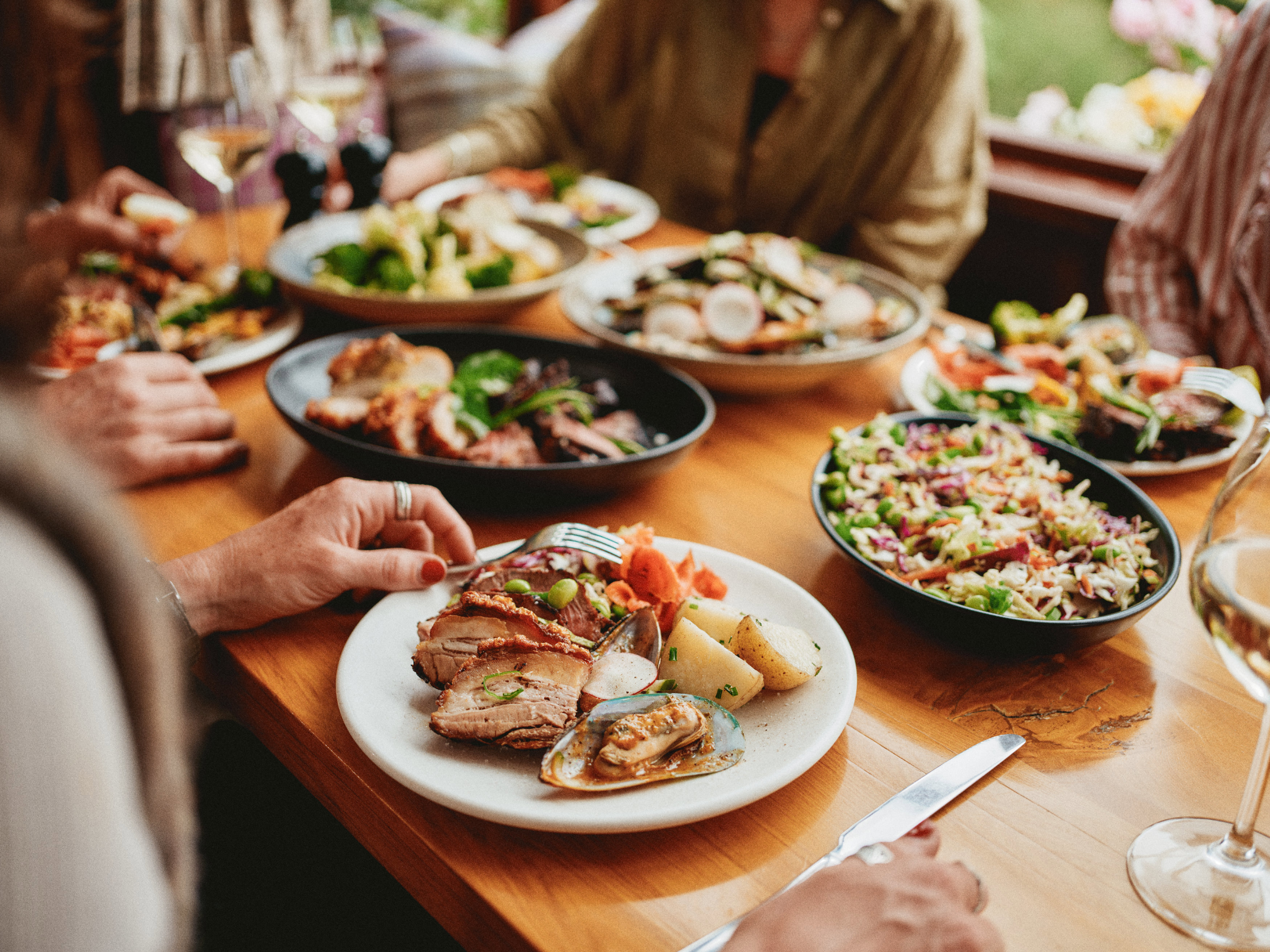 Guests sharing a spread of dishes at Walter Peak Gourmet BBQ, including sliced meat, mussels, fresh salads, and vegetables, with wine glasses on a warm timber table.