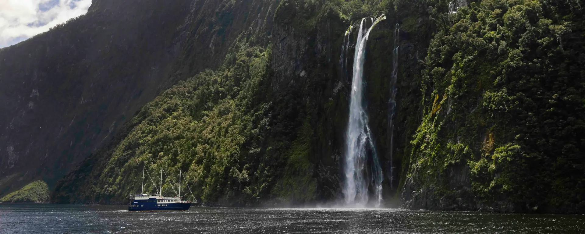 Cruise vessel passing waterfalls beneath steep Milford Sound mountains.