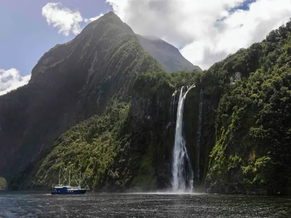 Cruise vessel passing waterfalls beneath steep Milford Sound mountains.