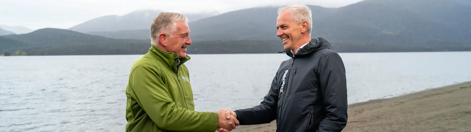 Two men, one in Department of Conservation uniform and one in RealNZ uniform, shake hands while standing on a beach.
