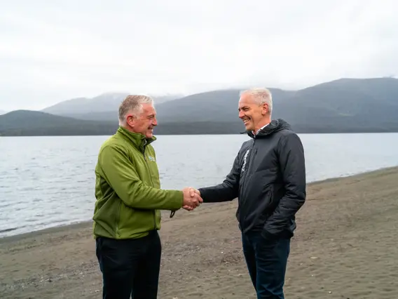 Two men, one in Department of Conservation uniform and one in RealNZ uniform, shake hands while standing on a beach.