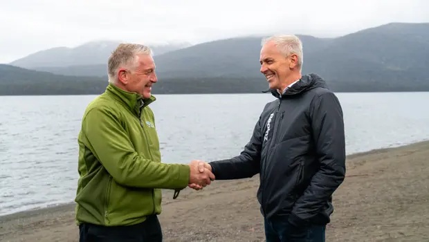 Two men, one in Department of Conservation uniform and one in RealNZ uniform, shake hands while standing on a beach.