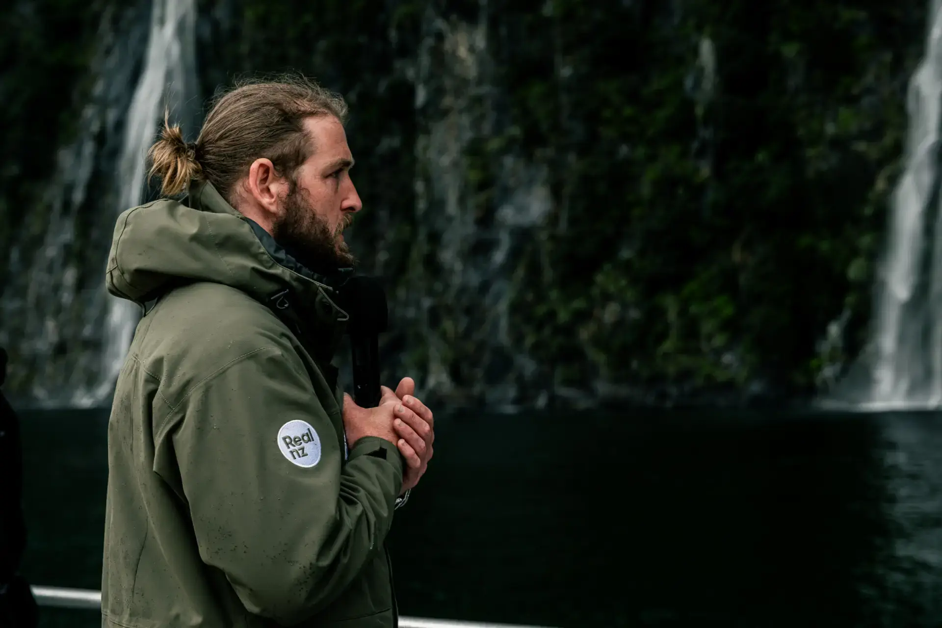 A man wearing a RealNZ branded rain jacket stands on an outside deck of a boat, speaking into a microphone with a waterfall in the background.