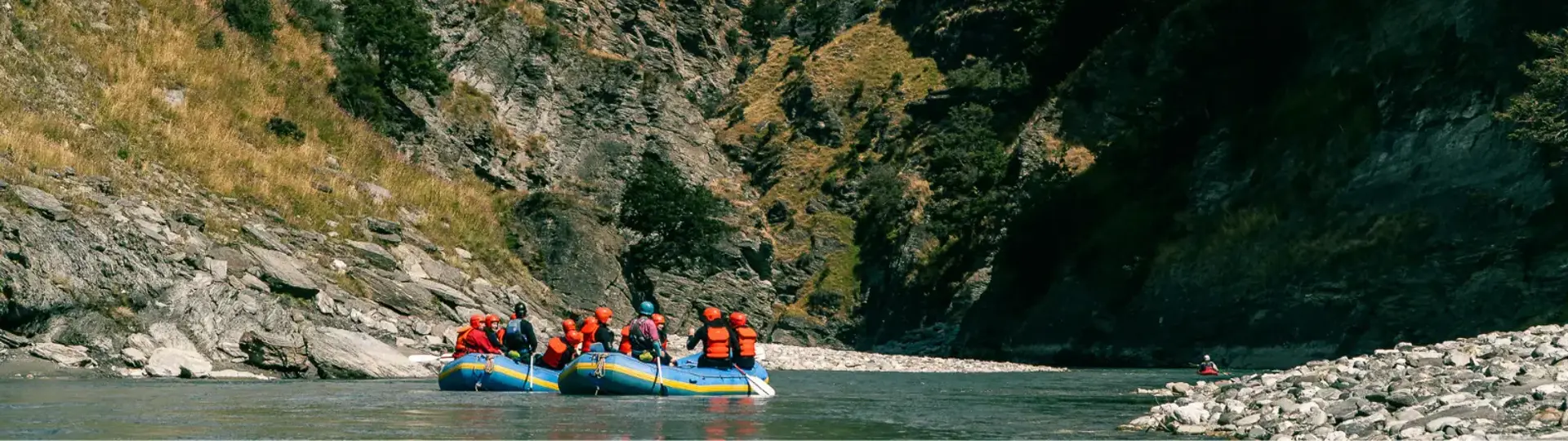Two whitewater rafting rafts, with teams of people on them wearing wetsuites, helmets and lifejackets, sit on still water surrounded by towering cliff faces. 