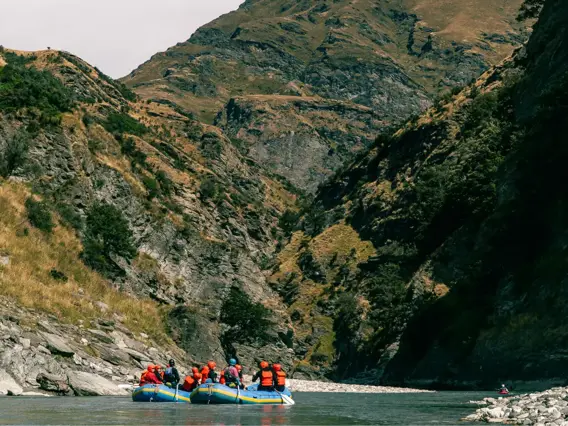 Two whitewater rafting rafts, with teams of people on them wearing wetsuites, helmets and lifejackets, sit on still water surrounded by towering cliff faces. 