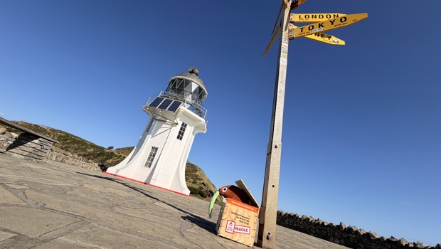 The famous lighthouse and sign at Cape Reigna, with a small soft toy brown kiwi in a translocation box sittng at the bottom of the sign. 