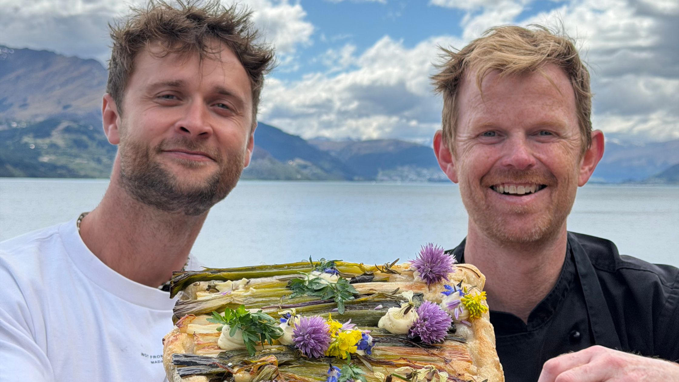 Two men smiling by the lake, holding a spring leek focaccia decorated with herbs and flowers.