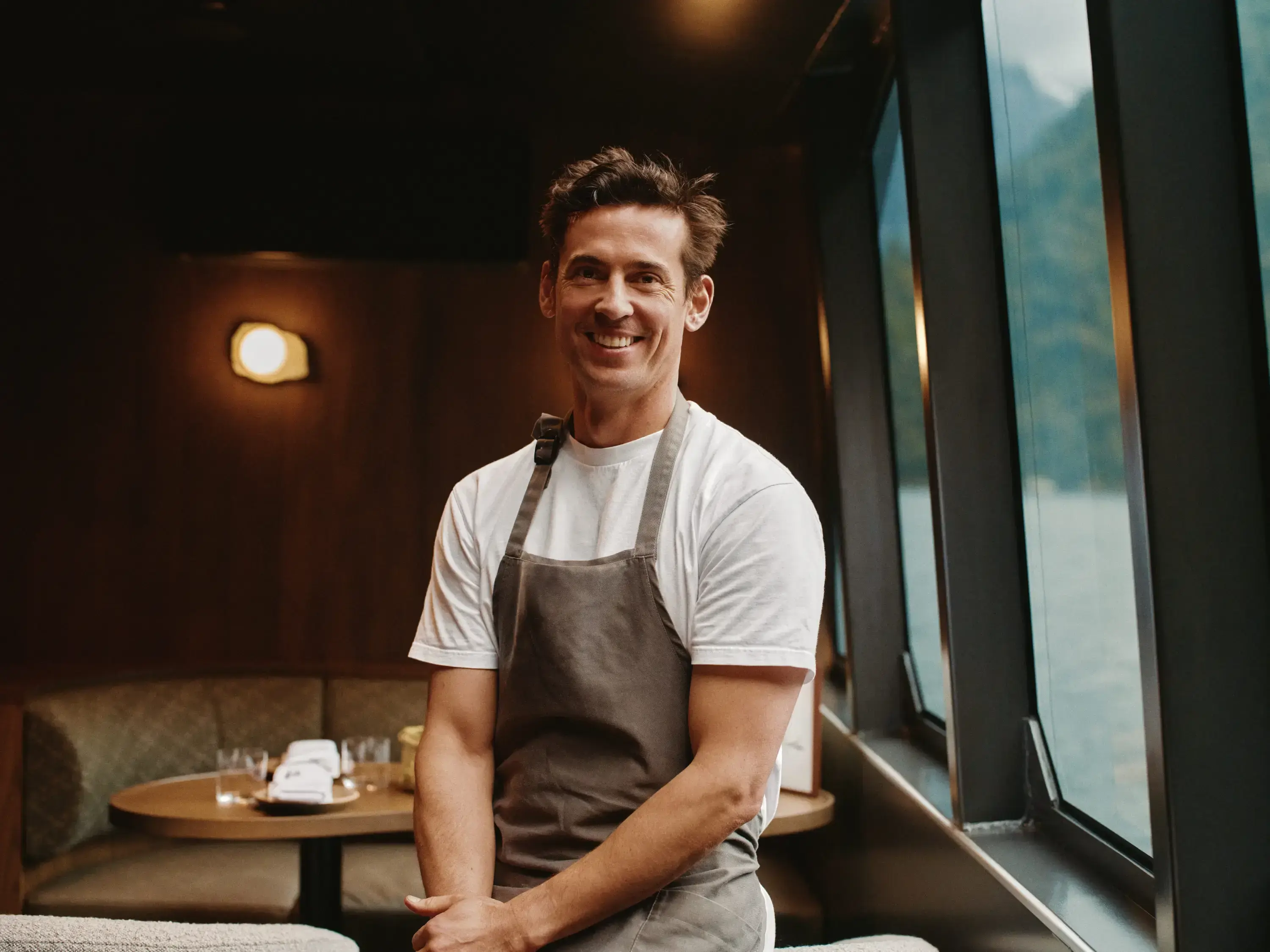 Male chef wearing white t-shirt and brown apron sits smiling in the modern dining area of MV Sinbad cruise vessel with dramatic Milford Sound fiordland landscape visible through large windows behind him.