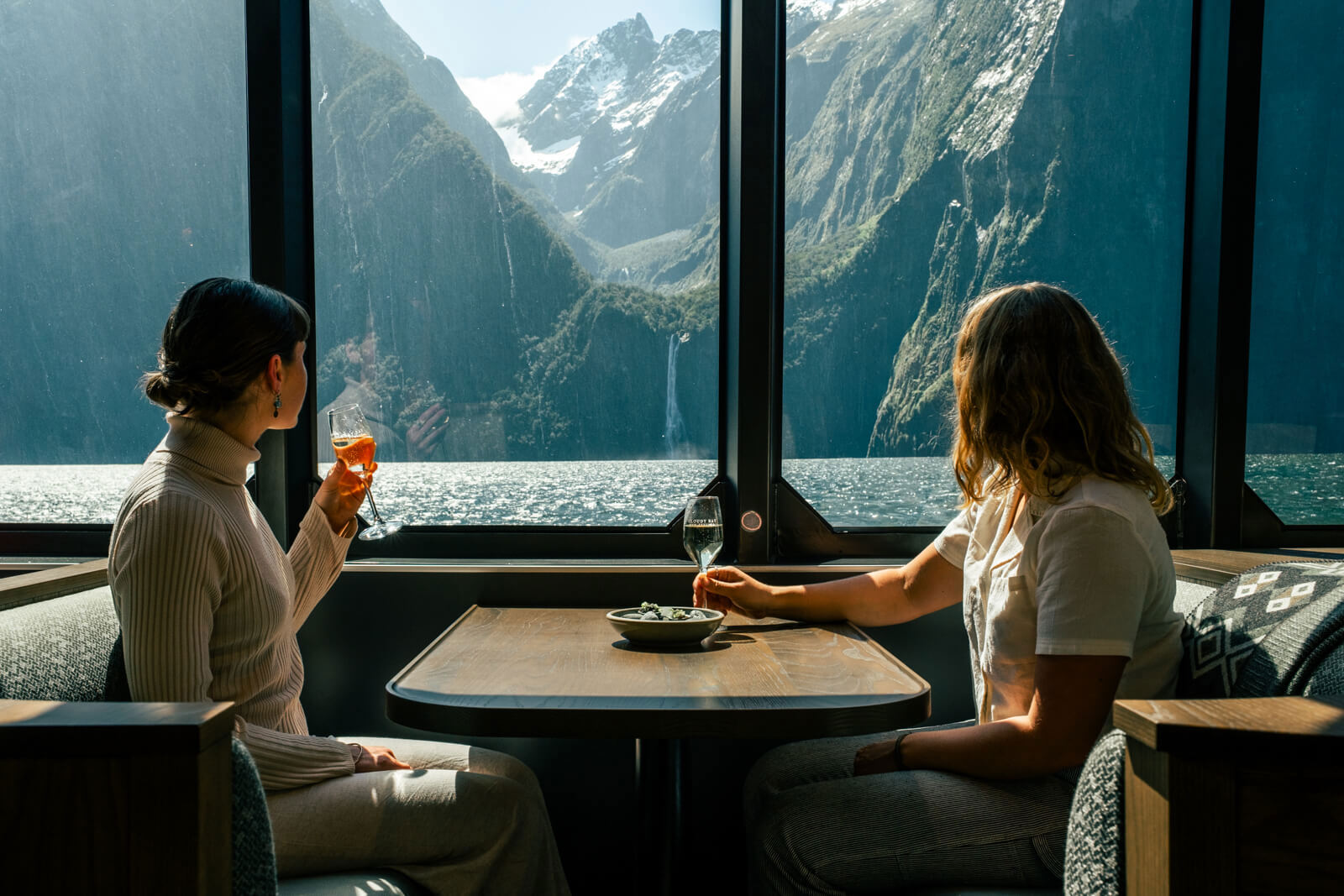 Two people dining with wine glasses at a table aboard a vessel, with a dramatic view of Milford Sound's steep fjord walls and a waterfall through large panoramic windows.