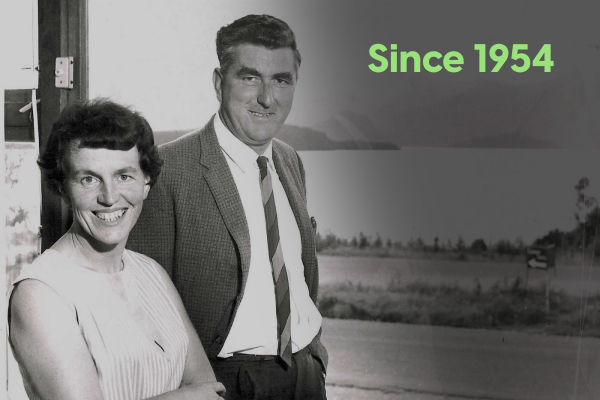 A black-and-white archival photo of a smiling couple, with a landscape in the background and green text reading "Since 1954."