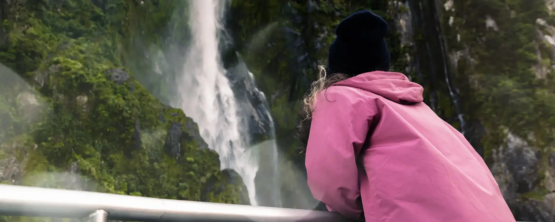 Guest in a pink jacket viewing a Milford Sound waterfall from the deck.